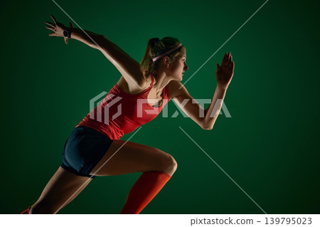 Close side profile of female sprinter accelerating on dark green studio background. 139795023