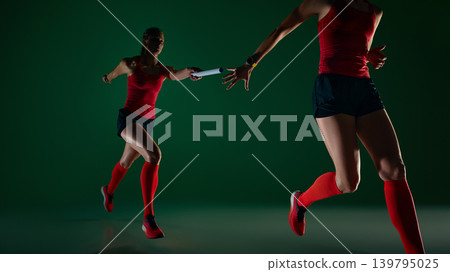 Two female runners passing relay baton in dramatic green studio scene. 139795025