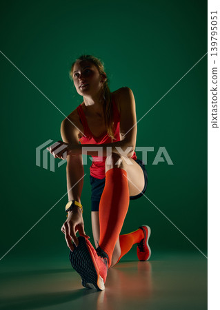 Female runner kneeling in deep stretch before training on green studio background. 139795051