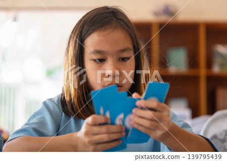Focused little girl holding and looking at blue domino pieces while playing a board game at home. 139795439