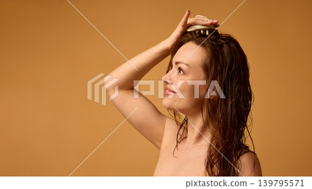 Smiling woman using scalp massager on wet brunette hair with orange copy space. 139795571