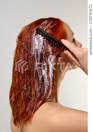 Woman combing red hair mask through wet hair during salon care routine. Woman combing red hair mask through wet hair during salon care routine. 139795672