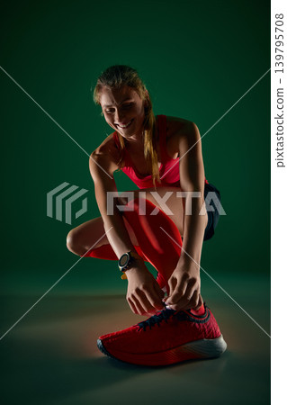 Smiling female athlete tying red running shoe before workout on green studio background. Smiling female athlete tying red running shoe before workout on green studio background. 139795708