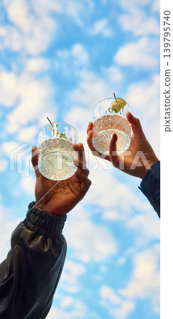 Two clear cocktails with cucumber garnish lifted toward blue cloudy sky. Two clear cocktails with cucumber garnish lifted toward blue cloudy sky. 139795740