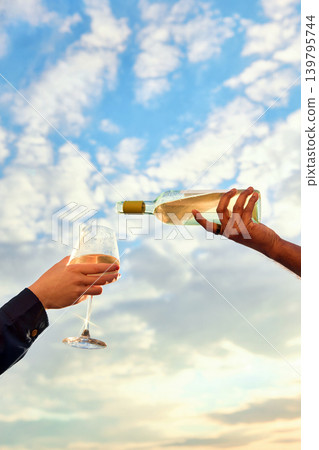 Hand pouring white wine into glass with cloudy blue sky background. Hand pouring white wine into glass with cloudy blue sky background. 139795744