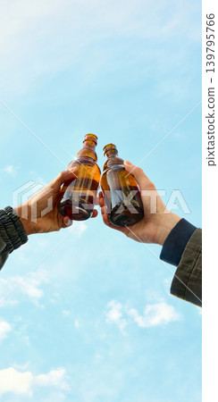 Two people clinking amber drink bottles under blue sky. 139795766