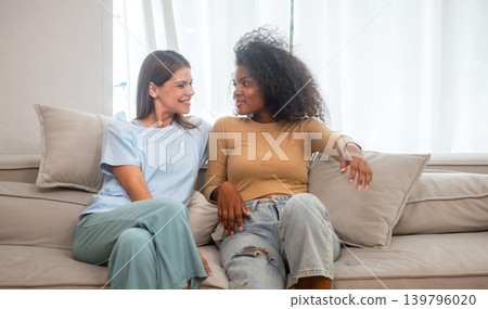Two women smile warmly while seated on a beige couch Two women smile warmly while seated on a beige couch 139796020