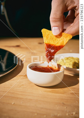 Hand holding tortilla chip coated with red salsa over dipping bowl in closeup Hand holding tortilla chip coated with red salsa over dipping bowl in closeup 139796123