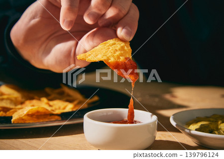Closeup hand dipping tortilla chip into red salsa above white bowl in warm sunlight Closeup hand dipping tortilla chip into red salsa above white bowl in warm sunlight 139796124