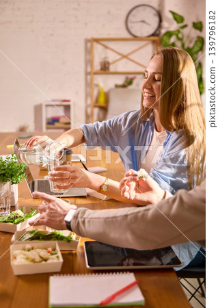 Smiling woman pouring water while having office lunch with coworkers. 139796182
