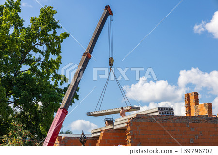 A crane lifts a large concrete slab to place it on a building site with walls of bricks and trees nearby on a sunny day A crane lifts a large concrete slab to place it on a building site with walls of bricks and trees nearby on a sunny day 139796702