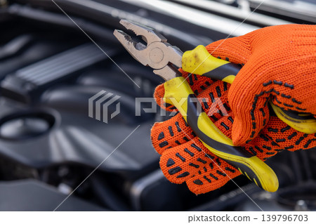 A man is working on a car engine in a garage, holding pliers and wearing bright gloves. He is focused on the task at hand A man is working on a car engine in a garage, holding pliers and wearing bright gloves. He is focused on the task at hand 139796703