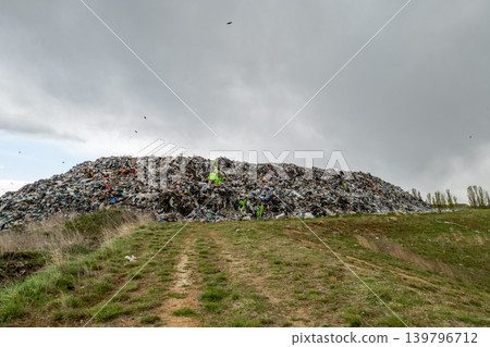 Workers are sorting through trash at a landfill site with a large mound of waste in the background and dark clouds overhead Workers are sorting through trash at a landfill site with a large mound of waste in the background and dark clouds overhead 139796712