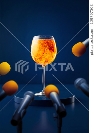 Bright orange cocktail with ice and citrus slice on podium with microphones on blue background. Bright orange cocktail with ice and citrus slice on podium with microphones on blue background. 139797508