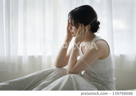 A young woman sitting on a bed, holding her head in her hands. A young woman sitting on a bed, holding her head in her hands. 139798086
