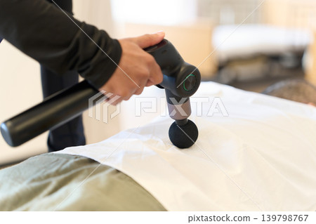 A man's hands as he massages his lower back with a massage gun. 139798767