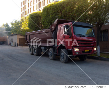 Red dump truck parked on a city street near modern buildings and green trees. Heavy industrial transport vehicle used for construction, cargo hauling, and urban infrastructure work Red dump truck parked on a city street near modern buildings and green trees. Heavy industrial transport vehicle used for construction, cargo hauling, and urban infrastructure work 139799096