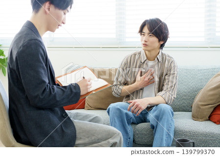 Counseling session: A young Japanese man sits on a sofa and talks, while a young Japanese doctor takes notes as he listens. Counseling session: A young Japanese man sits on a sofa and talks, while a young Japanese doctor takes notes as he listens. 139799370