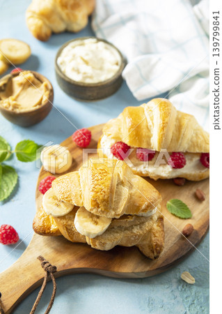 French pastry, freshly baked croissants with peanut butter, raspberries and banana on a blue stone background. Healthy food dessert concept. French pastry, freshly baked croissants with peanut butter, raspberries and banana on a blue stone background. Healthy food dessert concept. 139799841