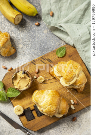 French pastry, freshly baked croissants with peanut butter and banana on a grey stone background. Healthy food dessert concept. View from above. French pastry, freshly baked croissants with peanut butter and banana on a grey stone background. Healthy food dessert concept. View from above. 139799990