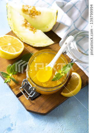 Sweet melon and citrus jam, homemade jelly preserves in small glass jar with fresh melon slices on a blue stone tabletop. Homemade preserve. Copy space. Sweet melon and citrus jam, homemade jelly preserves in small glass jar with fresh melon slices on a blue stone tabletop. Homemade preserve. Copy space. 139799994