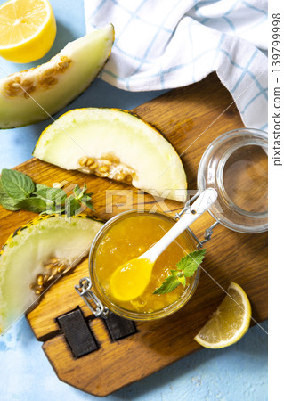 Sweet melon and citrus jam, homemade jelly preserves in small glass jar with fresh melon slices on a blue stone tabletop. Homemade preserve. View from above. Sweet melon and citrus jam, homemade jelly preserves in small glass jar with fresh melon slices on a blue stone tabletop. Homemade preserve. View from above. 139799998