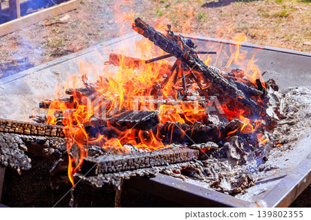 Bright flames rise from burning wood in metal fire pit under sun at grassy area with trees around Bright flames rise from burning wood in metal fire pit under sun at grassy area with trees around 139802355