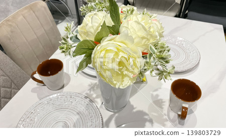 Cozy table setting with a white vase of artificial peonies and ceramic mugs on a marble surface 139803729