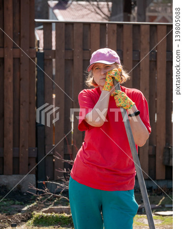 Mature woman gardener in red t-shirt leaning on garden tool in backyard. Caucasian lady in baseball cap working outdoors. Agriculture, vegetable garden maintenance and hobby lifestyle concept. Mature woman gardener in red t-shirt leaning on garden tool in backyard. Caucasian lady in baseball cap working outdoors. Agriculture, vegetable garden maintenance and hobby lifestyle concept. 139805088