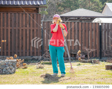 Woman gardener resting with rake in backyard. Mature lady cleaning lawn in spring season. Housework and yard maintenance. Gardening concept, lifestyle activity at home property. Woman gardener resting with rake in backyard. Mature lady cleaning lawn in spring season. Housework and yard maintenance. Gardening concept, lifestyle activity at home property. 139805090