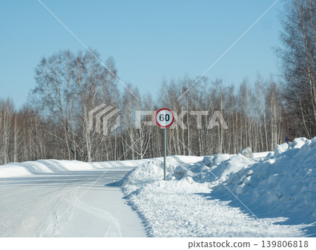 Speed limit sign 60 on a snowy rural road with forest in winter. Winter driving conditions concept with clear blue sky backdrop. Speed limit sign 60 on a snowy rural road with forest in winter. Winter driving conditions concept with clear blue sky backdrop. 139806818