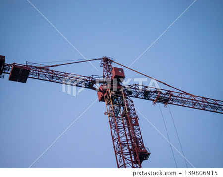 Red construction tower crane against clear blue sky outdoors. Industrial lifting equipment for building high-rise structures and urban development. 139806915