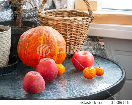 Pumpkin, apples, mandarins, and basket on a glass table. Holiday harvest still life composition. Autumn seasonal decor. 139806916