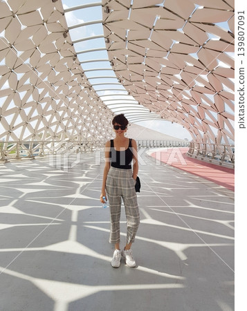 Woman posing on a modern architectural bridge with geometric metal structure. Stylish lady visiting city landmark for urban travel and tourism exploration. Fashionable summer lifestyle. Woman posing on a modern architectural bridge with geometric metal structure. Stylish lady visiting city landmark for urban travel and tourism exploration. Fashionable summer lifestyle. 139807091
