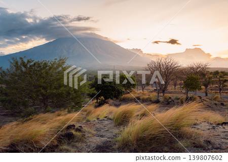Agung volcano landscape at sunset with dry grass, trees and dramatic cloudy sky Agung volcano landscape at sunset with dry grass, trees and dramatic cloudy sky 139807602