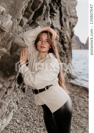 Woman cliff beach posing against a rocky coastal wall on a pebble shore during a calm overcast day Woman cliff beach posing against a rocky coastal wall on a pebble shore during a calm overcast day 139807647