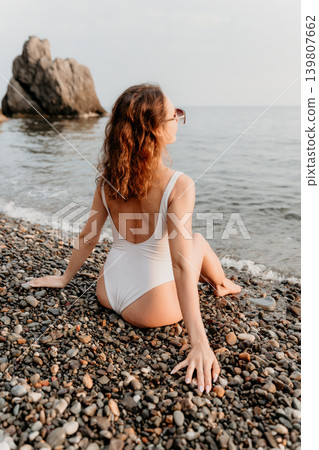 Woman beach swimwear relaxed brunette sits on a pebble shore looking at the sea and cliffs during summer vacation 139807662