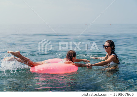 Family beach swimming mother and daughter enjoy a sunny summer vacation day playing together in the calm blue ocean with a bright pink inflatable ring Family beach swimming mother and daughter enjoy a sunny summer vacation day playing together in the calm blue ocean with a bright pink inflatable ring 139807666