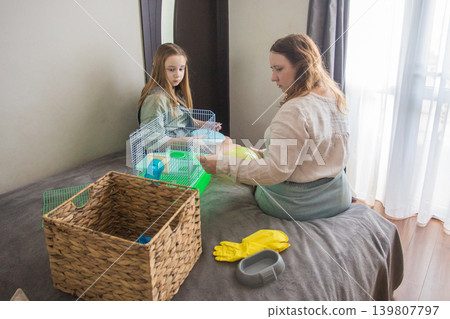 woman an girl sit on a bed in a room. Mom is holding a green hamster house with hamster 139807797