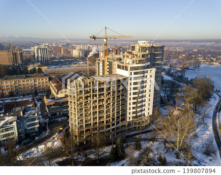 Apartment or office tall building under construction, aerial view. Tower crane silhouette Snowy field and distant city on bright blue sky copy space background. Drone photography. 139807894