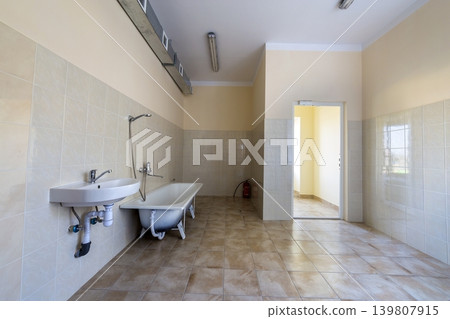 Interior of spacious light hospital or kindergarten bathroom with white bathtub, sink, air ventilation duct and tiled floor and walls. Interior of spacious light hospital or kindergarten bathroom with white bathtub, sink, air ventilation duct and tiled floor and walls. 139807915