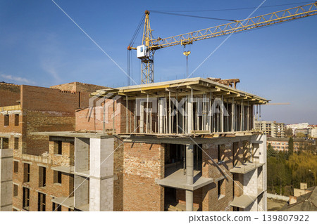 Apartment or office tall building under construction, top view. Brick walls, scaffolding and concrete support pillars. Tower crane on bright blue sky copy space background. Drone aerial photography. Apartment or office tall building under construction, top view. Brick walls, scaffolding and concrete support pillars. Tower crane on bright blue sky copy space background. Drone aerial photography. 139807922