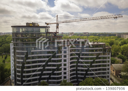Apartment or office tall building unfinished under construction among green tree tops. Tower cranes on bright blue sky copy space background. Apartment or office tall building unfinished under construction among green tree tops. Tower cranes on bright blue sky copy space background. 139807936