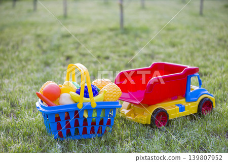 Car truckand a basket with toy fruit and vegetables. Bright plastic colorful toys for children outdoors on sunny summer day. Car truckand a basket with toy fruit and vegetables. Bright plastic colorful toys for children outdoors on sunny summer day. 139807952