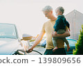 Young man holding baby and install cable plug to his electric car with home charging station. Residential EV Charging Station. Sustainable Power of a Wall-Mounted Charger. Future of Clean Transport. 139809656