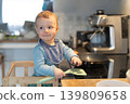 Baby boy standing on learning tower near induction stove with frying pan and holding a spatula in hand. Toddler repeating actions after parents, learning to cook food. Everyday kid development routine 139809658
