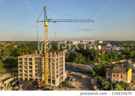 Apartment or office tall building under construction. Brick walls, glass windows, scaffolding and concrete support pillars. Tower crane on bright blue sky copy space background. Apartment or office tall building under construction. Brick walls, glass windows, scaffolding and concrete support pillars. Tower crane on bright blue sky copy space background. 139810870