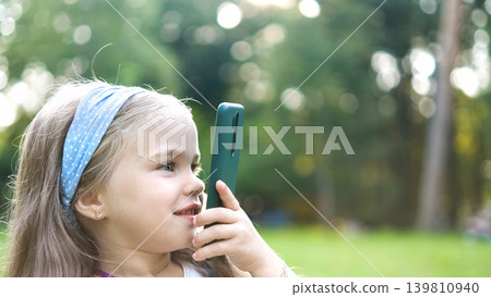 Happy little child girl having conversation talking on her cellphone in summer park. Happy little child girl having conversation talking on her cellphone in summer park. 139810940