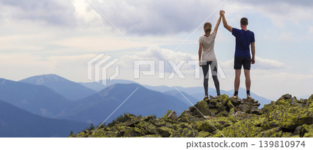 Back view of young tourist couple, athletic man and slim girl standing with raised arms holding hands on rocky mountain top enjoying fantastic panorama. Tourism, traveling and climbing concept. Back view of young tourist couple, athletic man and slim girl standing with raised arms holding hands on rocky mountain top enjoying fantastic panorama. Tourism, traveling and climbing concept. 139810974