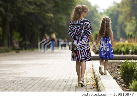 Back view of young blond long-haired attractive woman and small child girl holding hands outdoors. Back view of young blond long-haired attractive woman and small child girl holding hands outdoors. 139810977